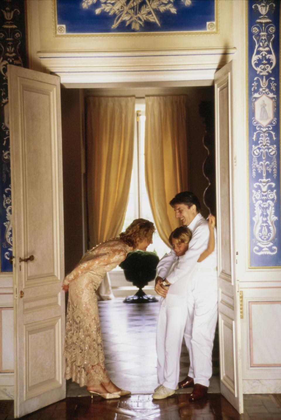 Fashion designer Emanuel Ungaro stands in the ballroom doorway, embracing his daughter, Cosima, while his wife Laura Emanuel (nee Bernabei) stands opposite, leaning forward towards Cosima, in their newly renovated Left Bank, Paris, France home with painted blue and white wall panels. Emanuel and Cosima are wearing white shirts and white pants, and Laura is wearing a long sleeve ankle length button-up back cream shimmer lace dress by Ungaro. Hair by Kerry Warn at John Frieda using John Frieda products. Makeup by Alan Milroy for Faces.