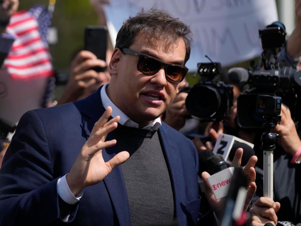 George Santos, who is wearing a blue blazer and sunglasses, speaking to a group of reporters with his hand raised for emphasis.