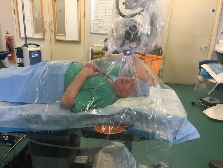 A man in surgical srubs lies on an operating table underneath a transparent plastic tent.