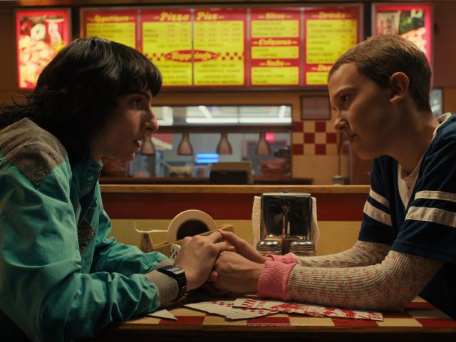 mike and eleven sitting together at a pizza restaurant, holding hands across a table and looking intently at the other in stranger things
