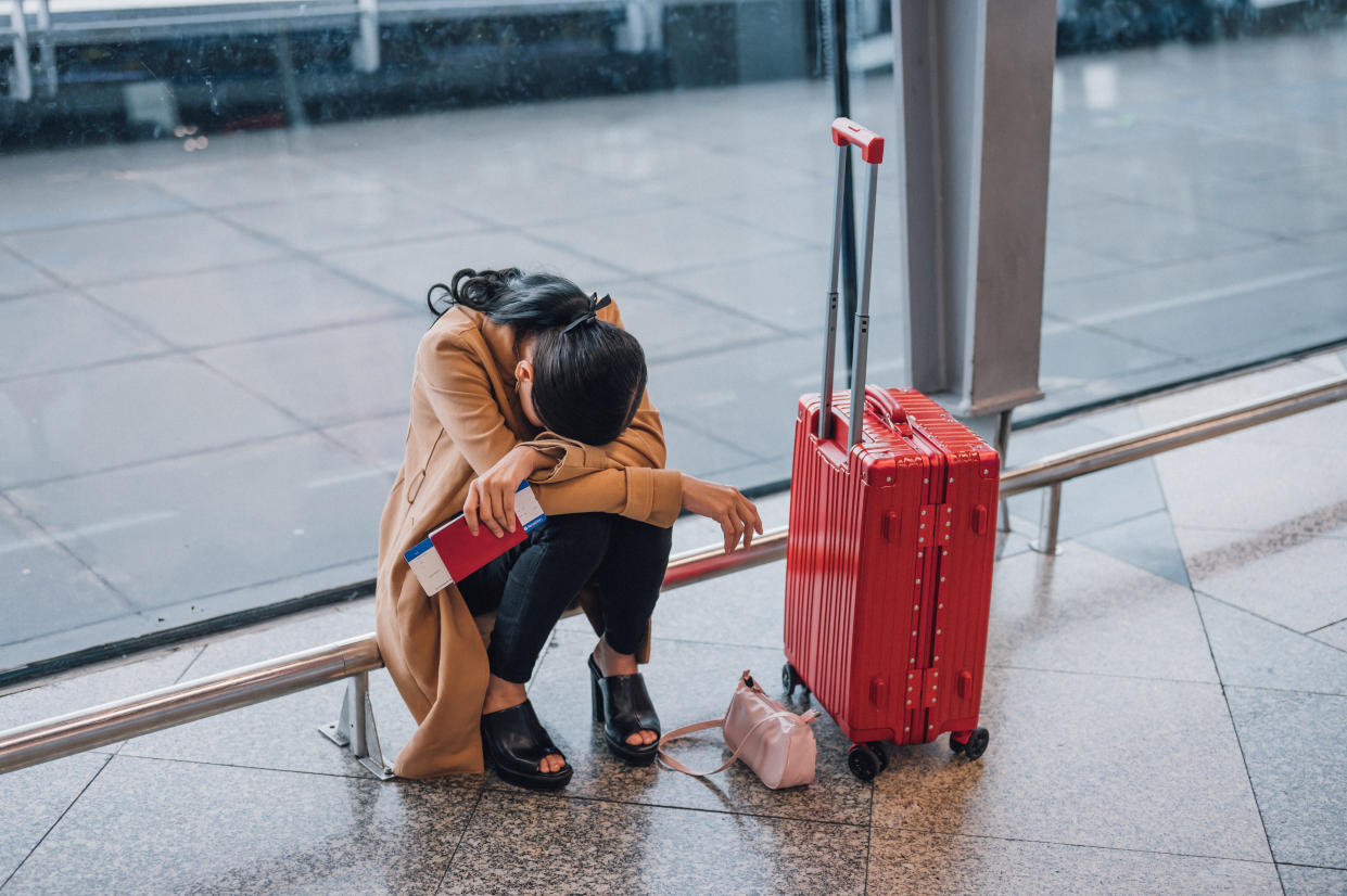 A forlorn traveler with ticket in hand and head resting on her crossed arms, with red rolling suitcase at her left.