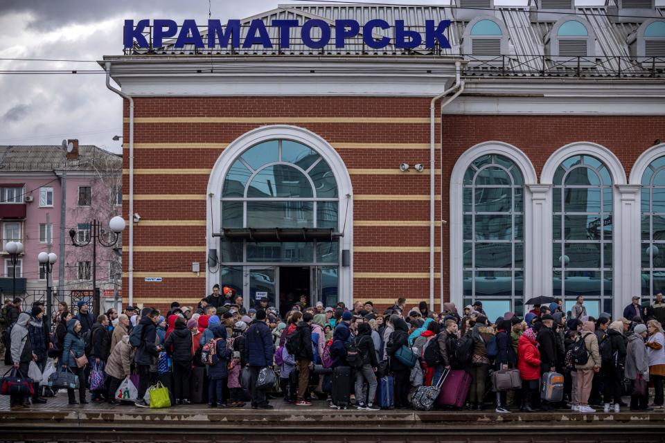 Dozens of people in cold weather clothes stand packed on an outdoor train station platform.