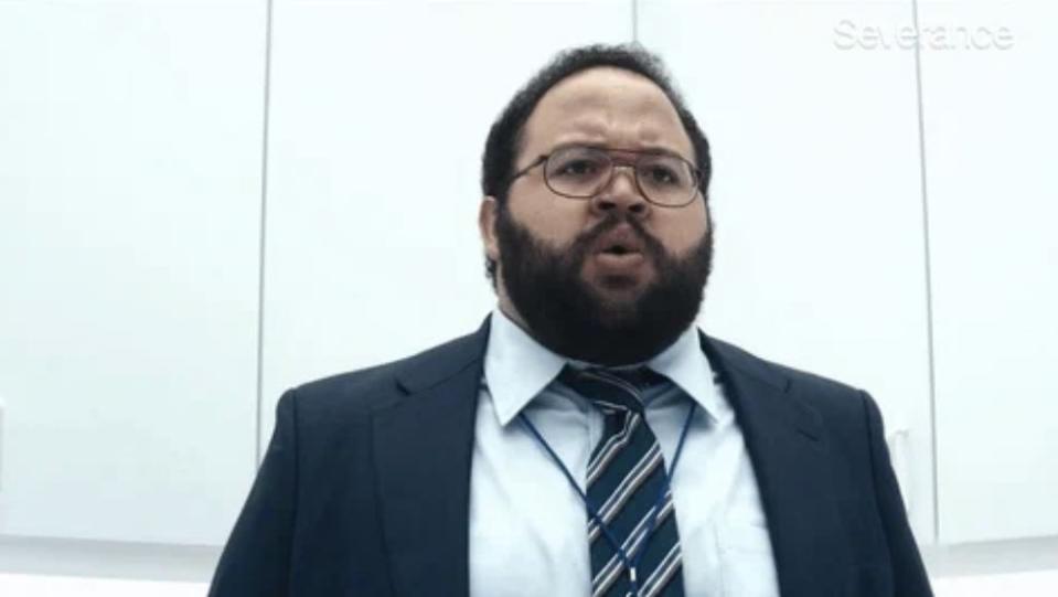 A man dressed in a suit and tie, appears shocked, standing in a brightly lit room with white cabinets behind him