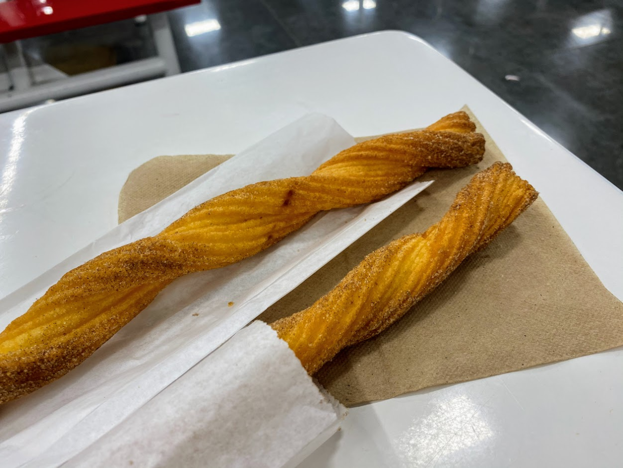 Two churros at the Sam's Club cafe, left one on top of the white bag it's served in and the right one still in the white bag, on a brown napkin, on a white table, grey concrete floor in the top and right background