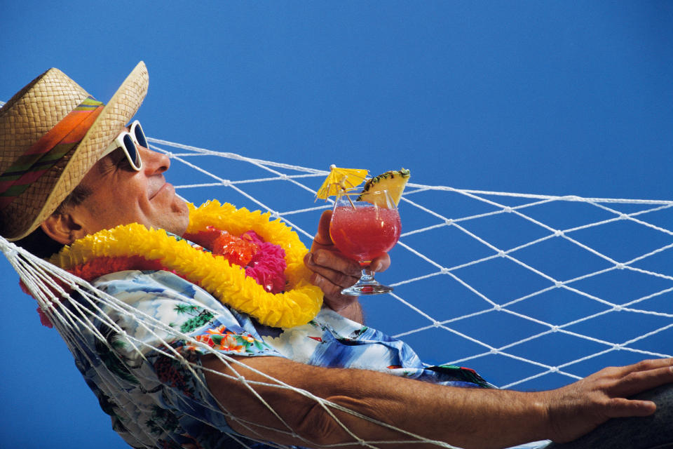 A middle aged man lies in a hammock with a tropical drink and sunglasses relaxing.