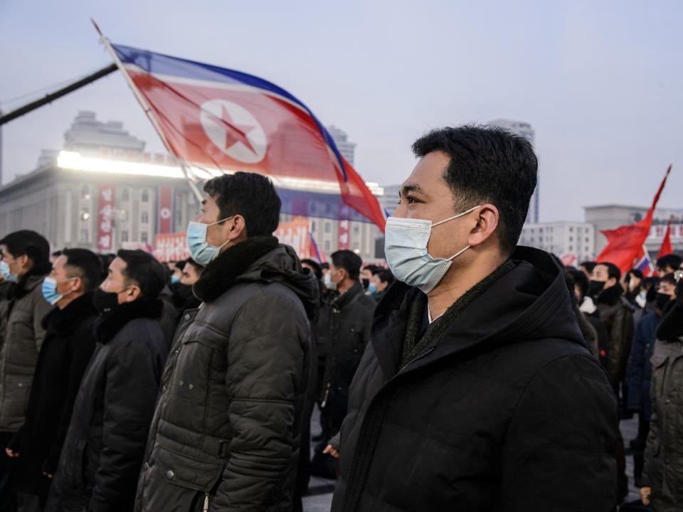 Masked North Korean people outdoors with the country's flag flying in the background.