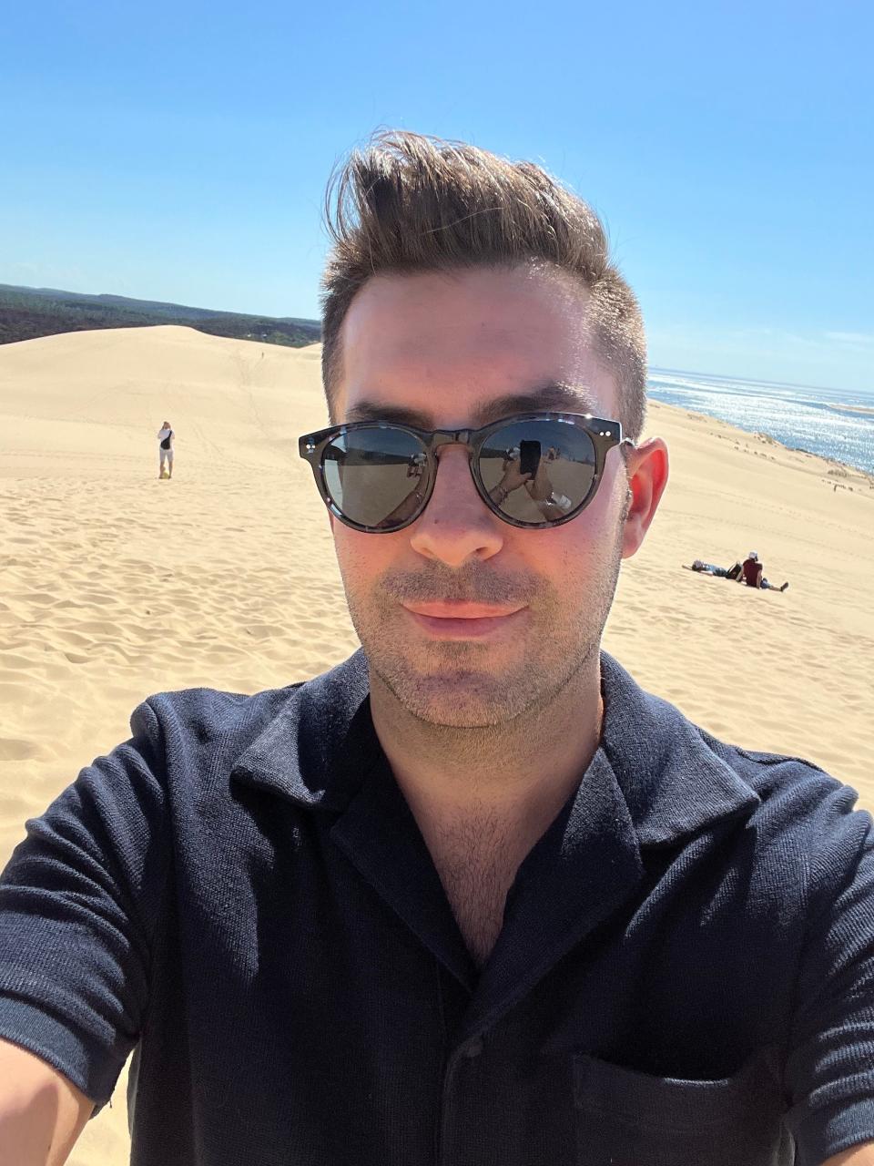 A man wearing sunglasses takes a selfie on the beach.