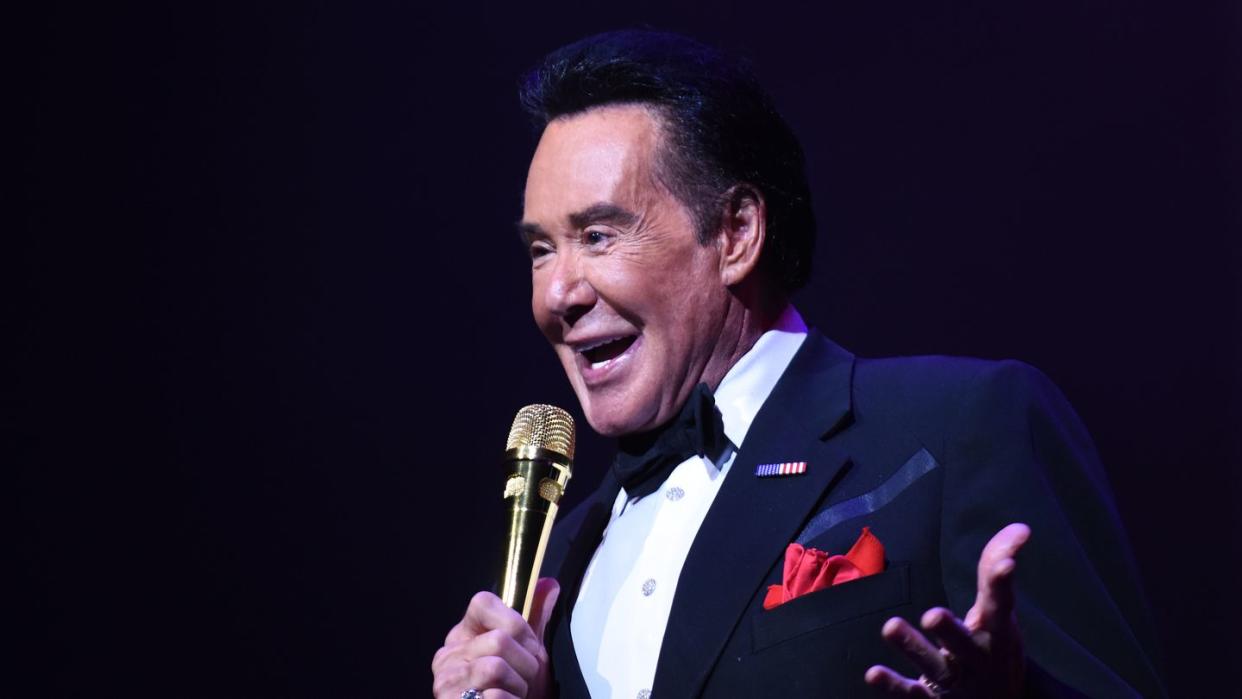 wayne newton smiles with his mouth open and holds a gold microphone to his face, his other hand is extended to the side, he wears a dark suit jacket, bow tie, white collared shirt and red pocket square