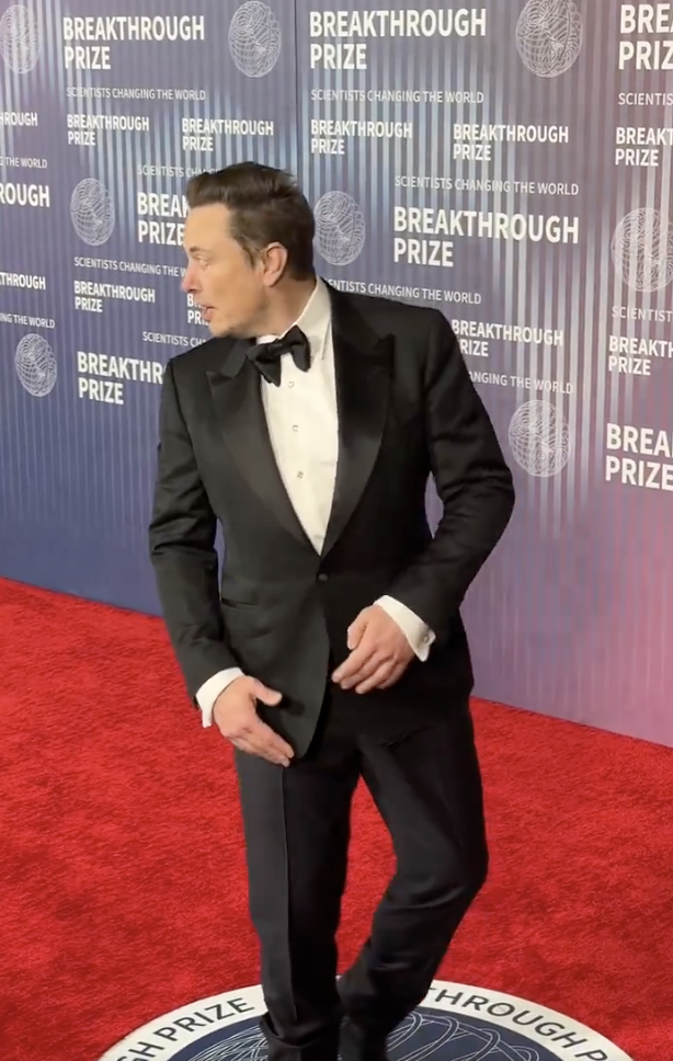 Elon Musk at an event in a black tuxedo with a bow tie, standing on a red carpet