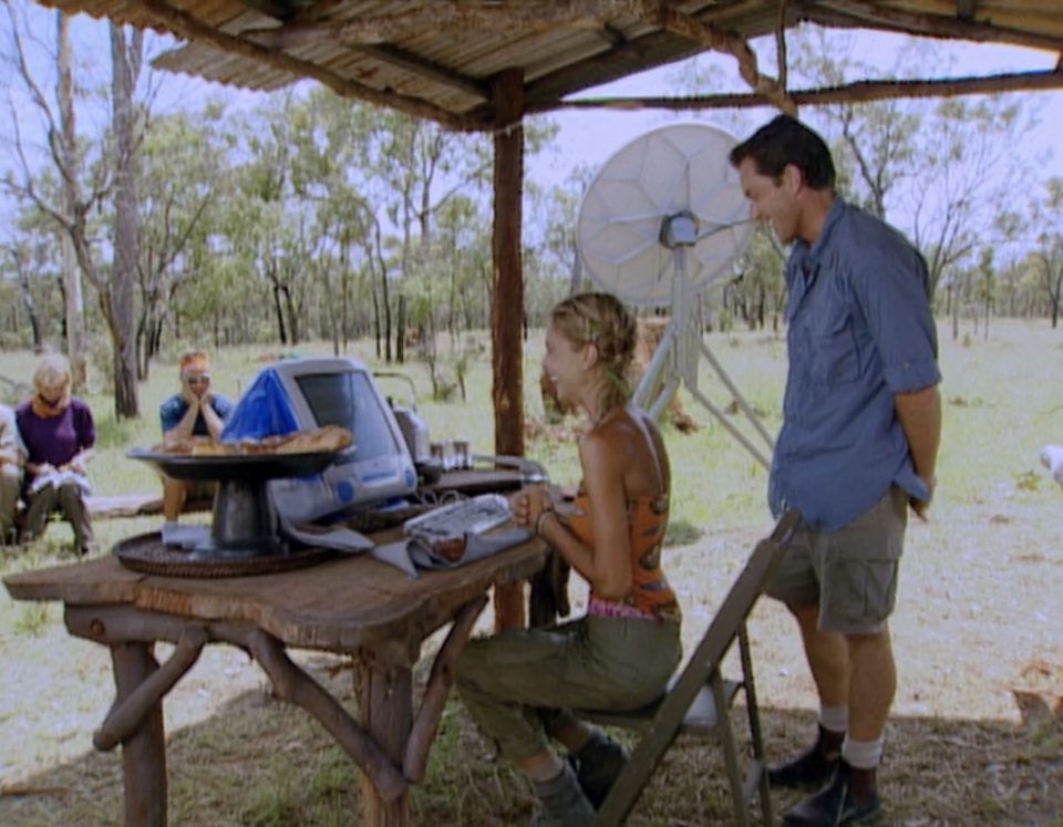 Elisabeth Filarski sits at a computer desk in the wilderness with Jeff Probst behind her on Survivor: The Australian Outback