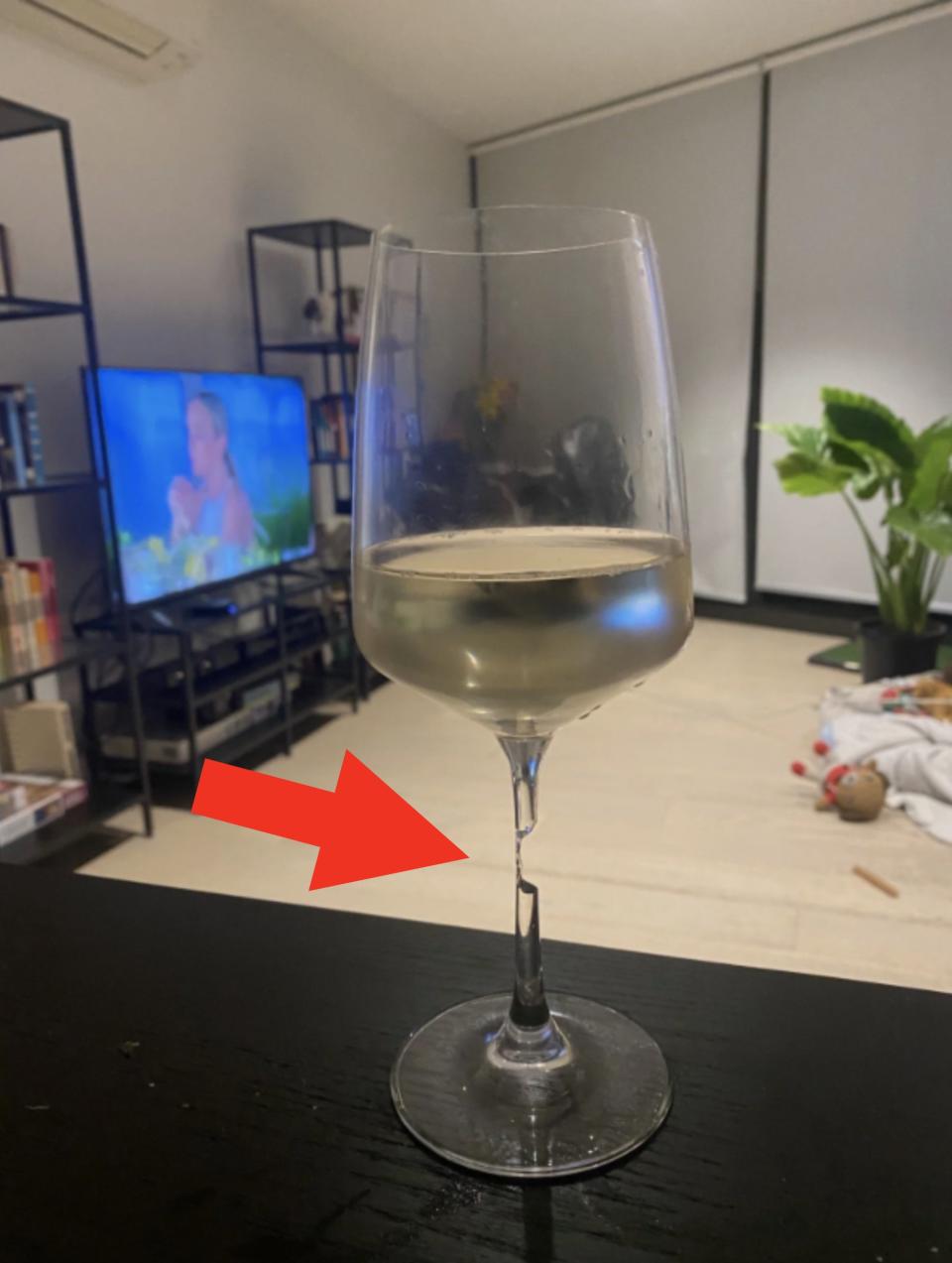 A glass of white wine on a table, with a TV showing a person in the background, modern living room with plants and bookshelves