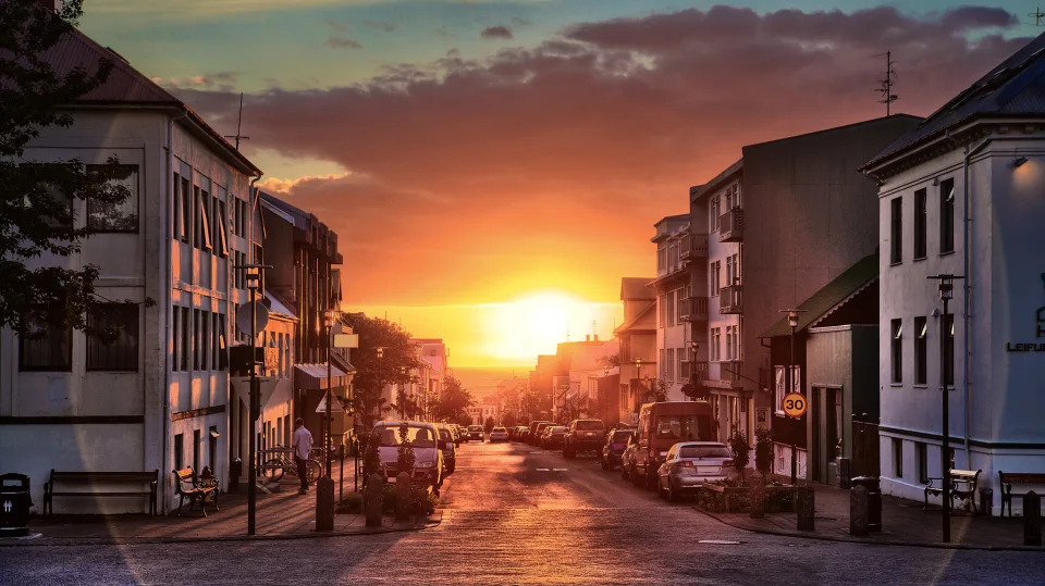 a street lined both sides with houses and buildings with the orange yellow bright glow of a setting sun in the distance.