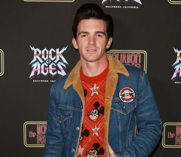 Closeup of Drake Bell on the red carpet wearing a denim jacket and a sweater with a Mickey Mouse motif