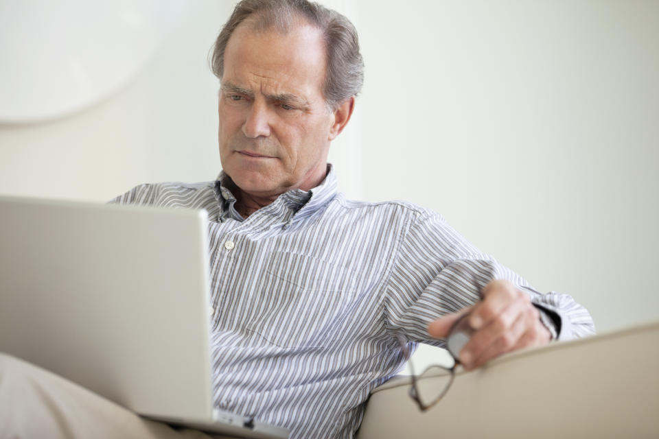 A mature man closely reading material on his laptop.