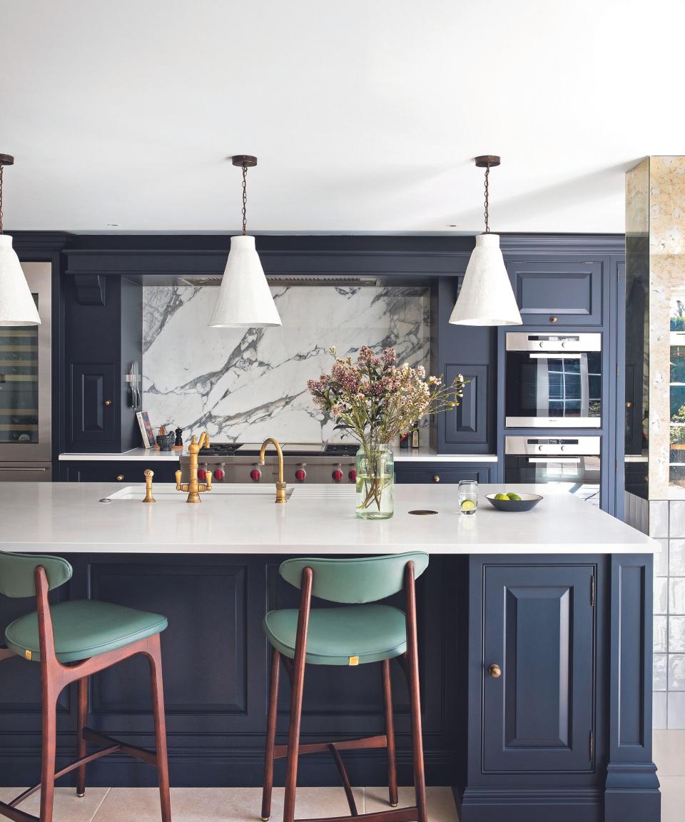 Open plan blue fitted kitchen with white worktops, kitchen island and green mid-century bar stools. Marble splashback.