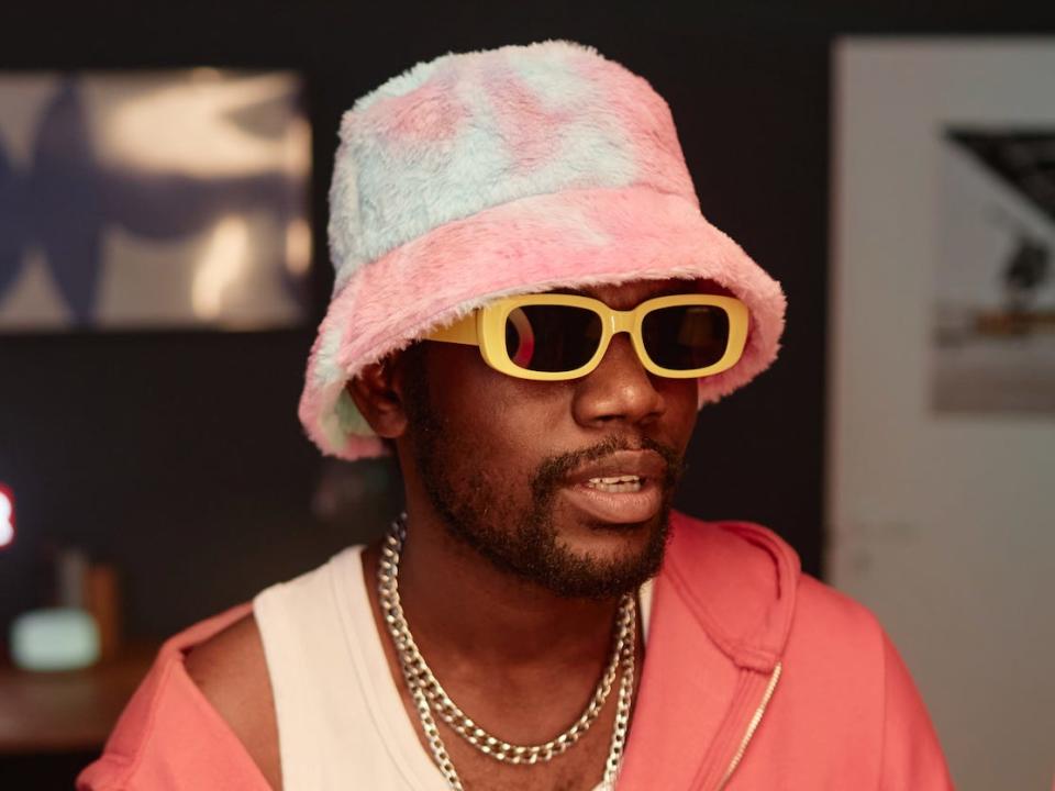 guy wearing a pink and blue fuzzy bucket hat, yellow sunglasses, a white tank top, and a salmon sweatshirt