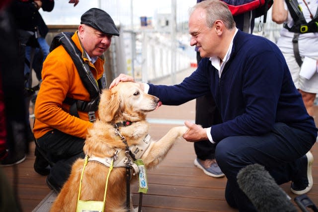 Ed Davey holds the paw of a guide dog as he pats its head watched by a man in an orange jacket and flat cap
