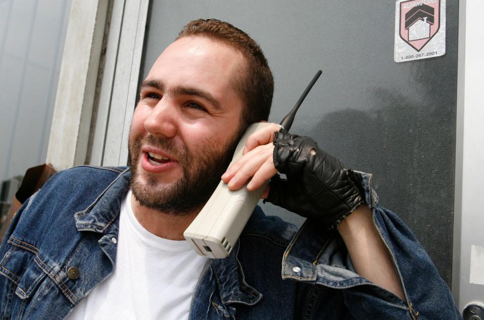 a man wearing a white tshirt, jean jacket, and black fingerless glove, talks on a white brick phone