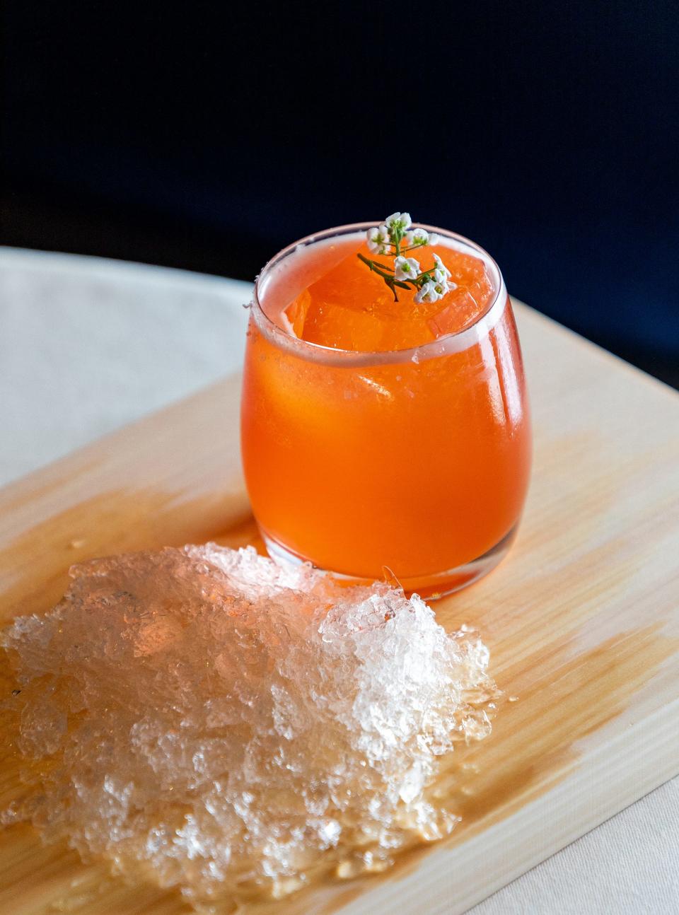 Orange beverage from Providence with white floral garnish on top of a wooden surface, with crushed ice on the side