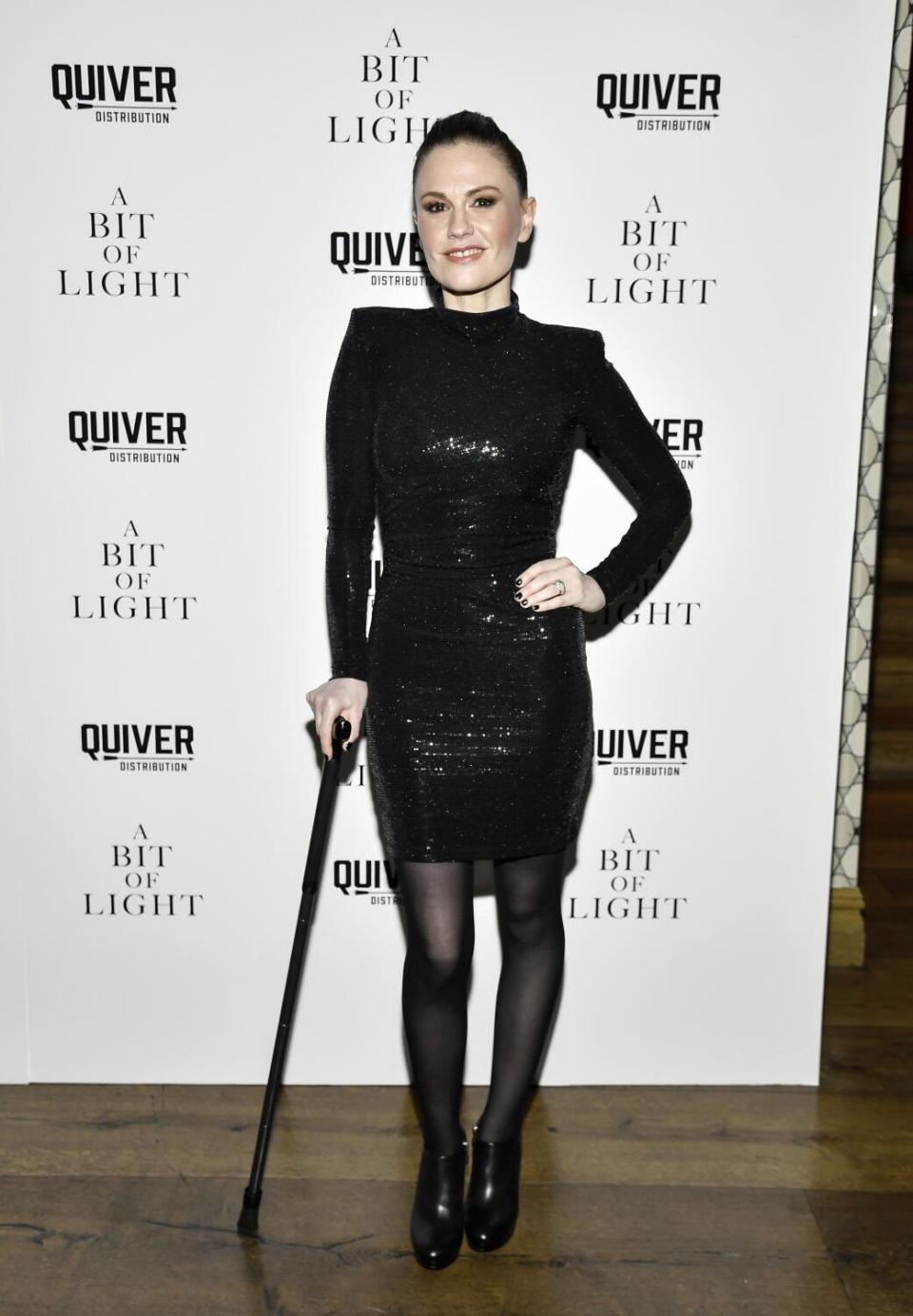 Anna Paquin in a black dress posing in front of a white backdrop, with one hand on her hip and the other holding a cane