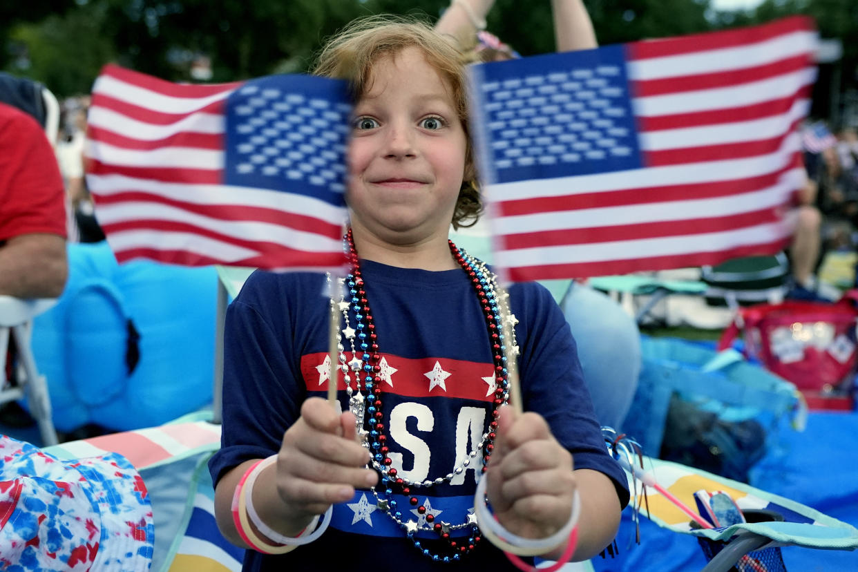 A child wearing a USA T-shirt, red-white-and-blue beads and glow-in-the-dark bracelets waves two American flags.