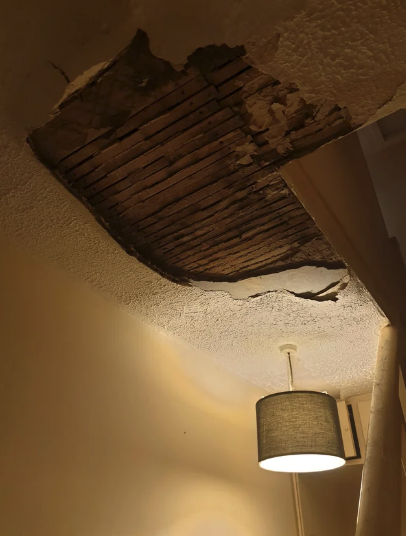 Ceiling with a large exposed hole showing broken plaster and wooden laths above a lit pendant lamp. The wall appears damaged