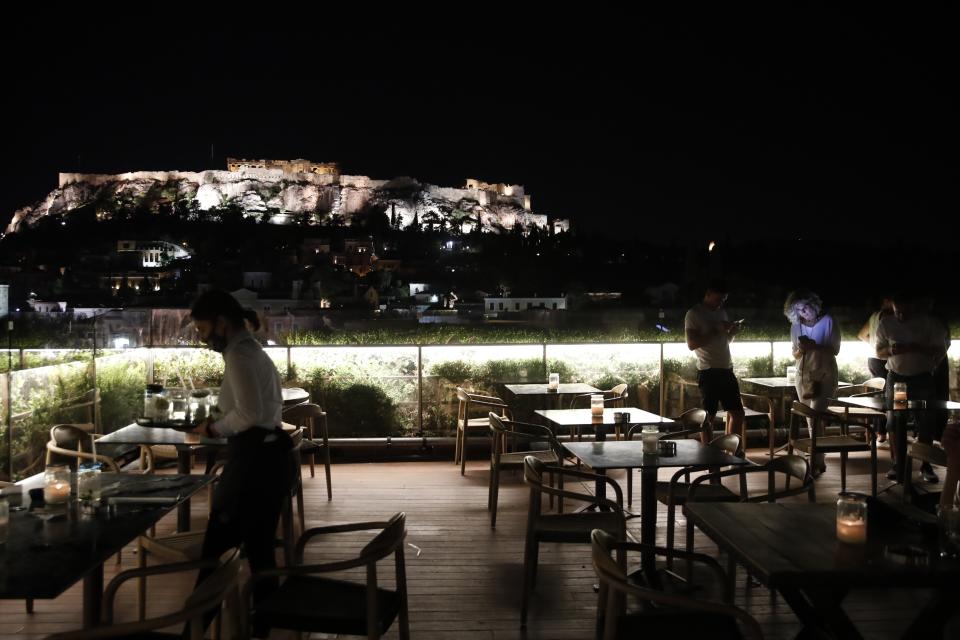 A waitress picks the drinks from a table as the last customers prepare to leave during the midnight closure of a bar with the Acropolis hill in the background in Athens, Friday, Aug. 21, 2020. Authorities in Greece are using free on-the-spot tests for ferry passengers and nightlife curfews on popular islands to stem a resurgence of the coronavirus after the country managed to dodge the worst of the pandemic. The number of confirmed virus cases and deaths in Greece remains lower than in many other European countries. (AP Photo/Thanassis Stavrakis)