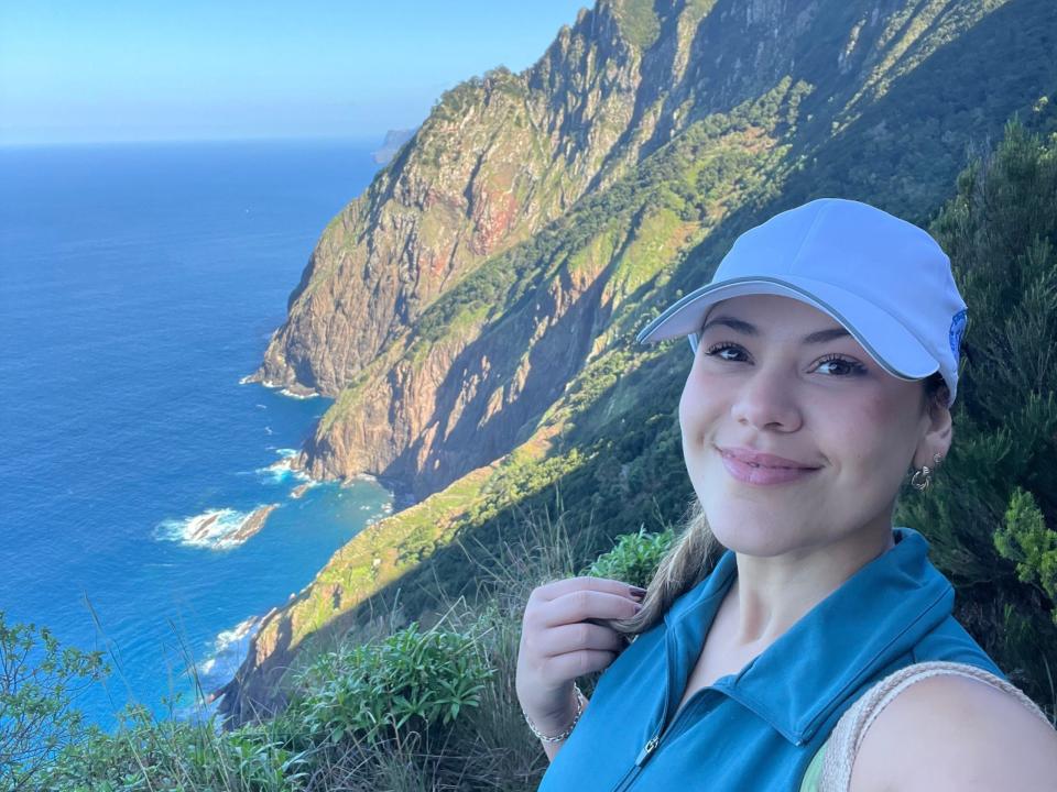 Victoria, die einen weißen Hut und ein blaues Oberteil trägt, posiert vor den Bergen am Wasser.