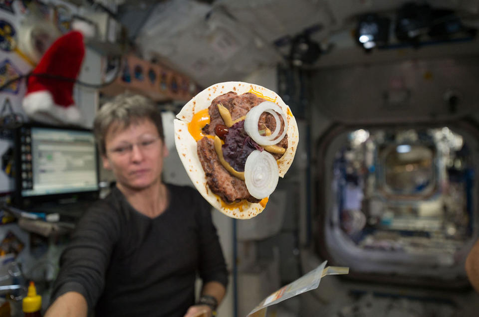 A "space cheeseburger," made using a tortilla for bread, is seen on board the International Space Station in late 2016. NASA astronaut Peggy Whitson floats in the background. <cite>NASA</cite>