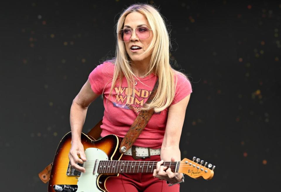 Sheryl Crow on stage playing a Fender guitar, wearing pink sunglasses and a pink T shirt with the slogan “Wonder Woman”