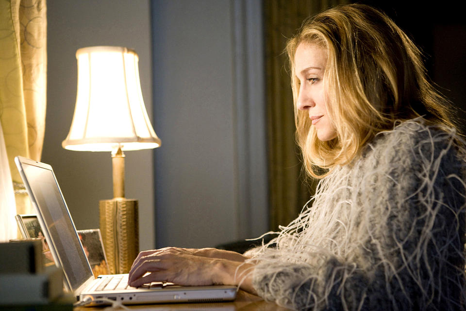 Sarah Jessica Parker typing on a laptop, wearing a textured, feather-like top, beside a lit lamp