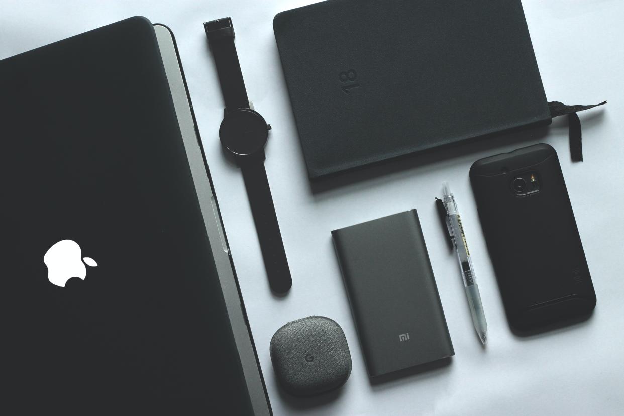  An Apple laptop, smart watch, Samsung phone, pen and notebook, all in black, laid out neatly across a white table. 