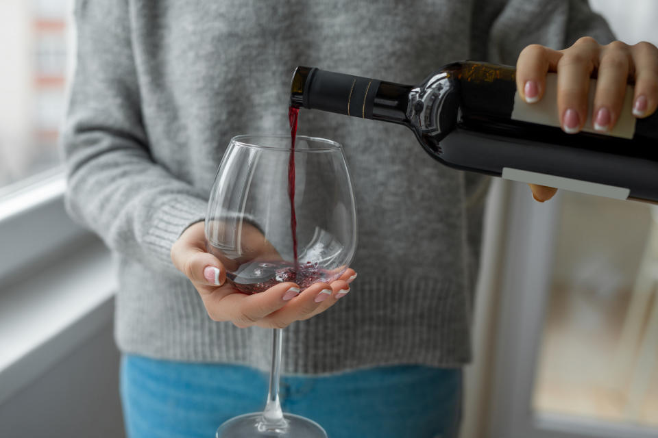 A person pours red wine from a bottle into a large wine glass indoors. The person is wearing a grey sweater and jeans