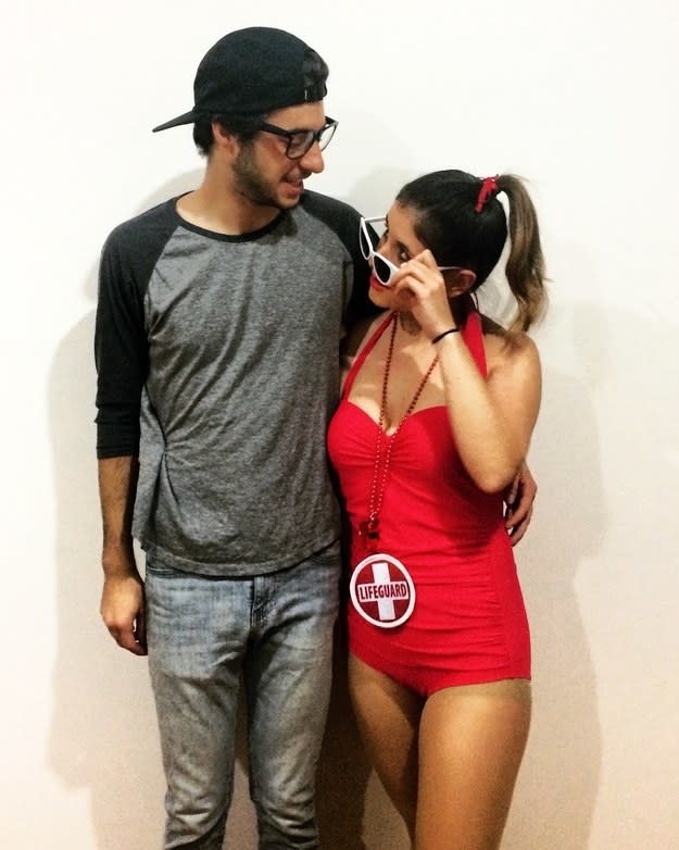 Man wearing a backward cap, T-shirt, and jeans hugging a woman in a "Lifeguard" bathing suit