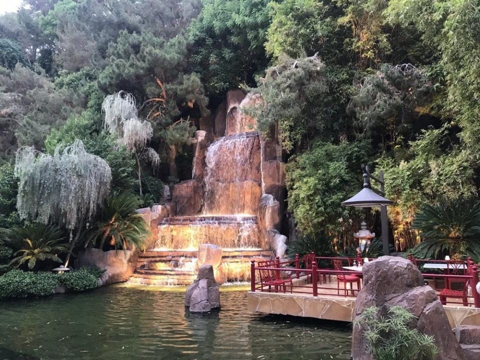 A waterfall with lush green trees on each side leading into a lake next to a seating area with red fencing.