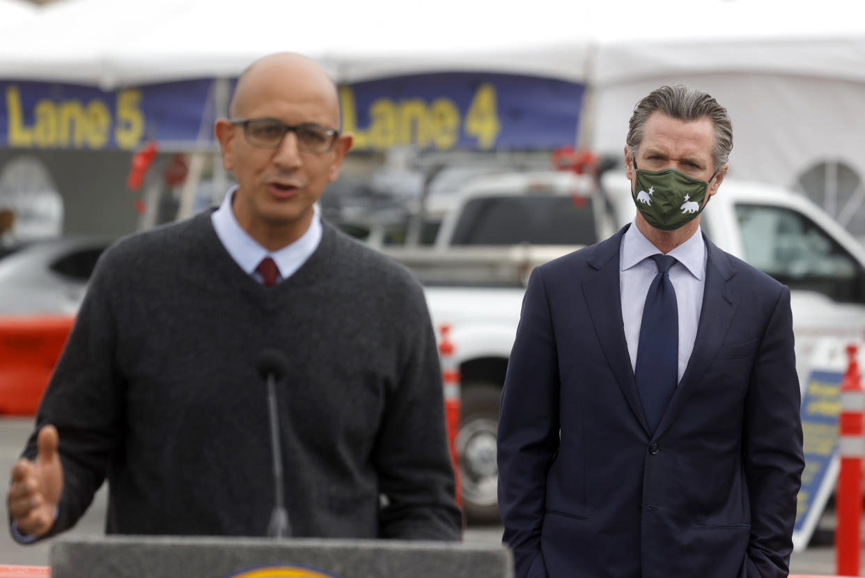 California Gov. Gavin Newsom looks on as California Health Secretary Mark Ghaly speaks into a microphone near cars and orange cones