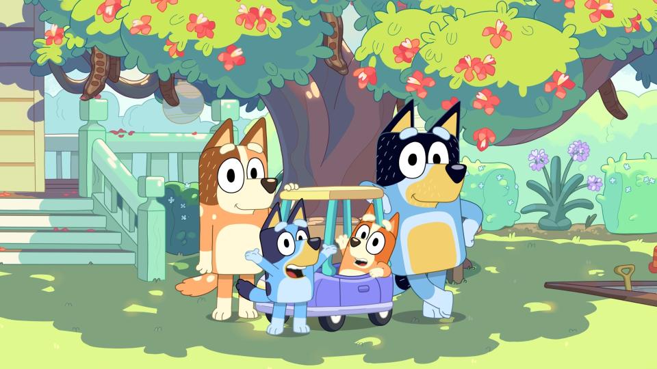 Animated canine family from "Bluey" with parents and children near a toy wagon under a tree