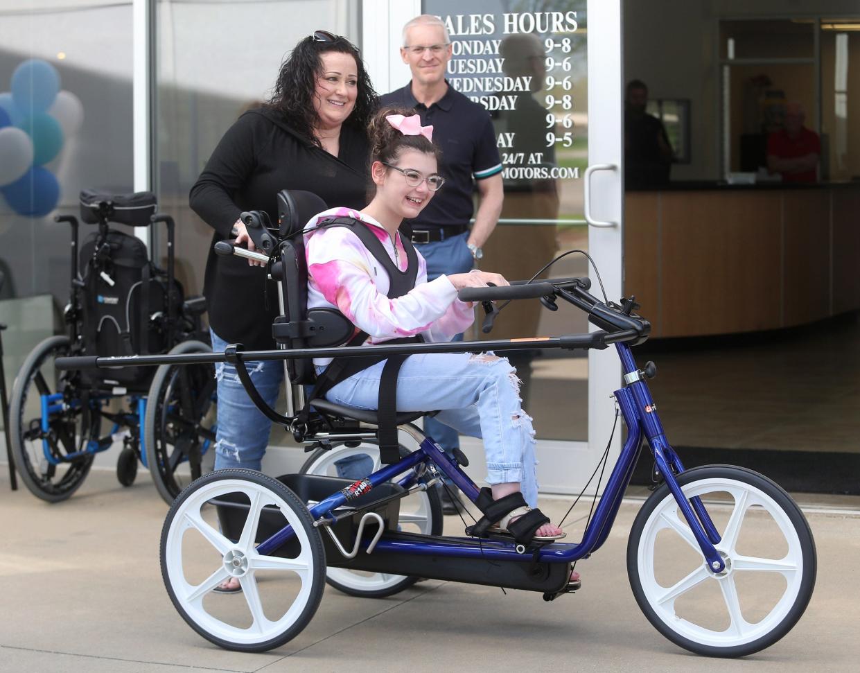 Bre McClure, left, helps her daughter Miah with her new adaptive tricycle that was presented this week to the Hoover High School freshman at Cain Toyota-BMW in Jackson Township. Looking on is dealership president Brian Cain.