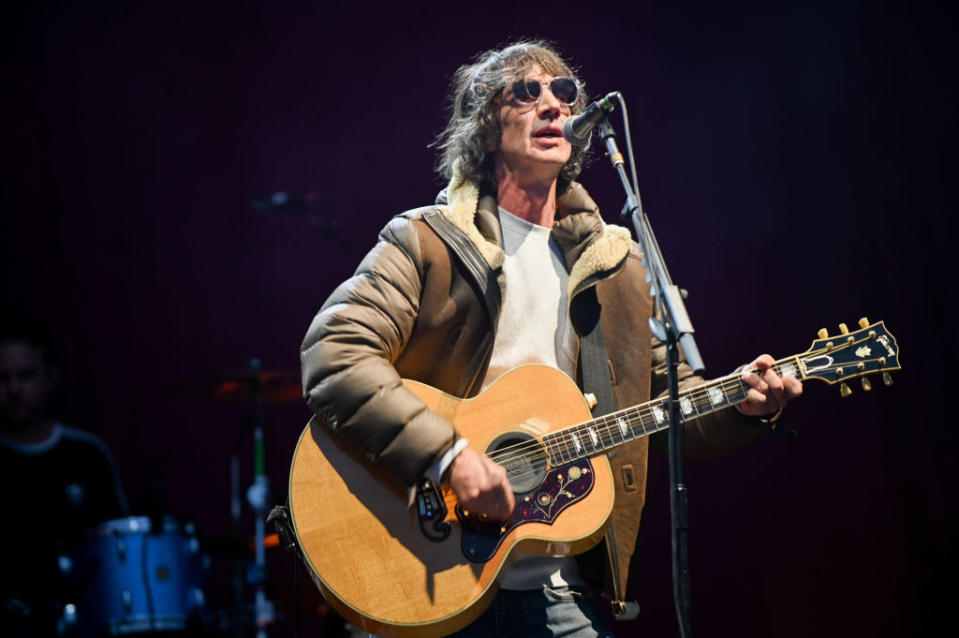 Richard Ashcroft performs on stage, wearing a puffy jacket and sunglasses, while playing an acoustic guitar. A drummer is visible in the background