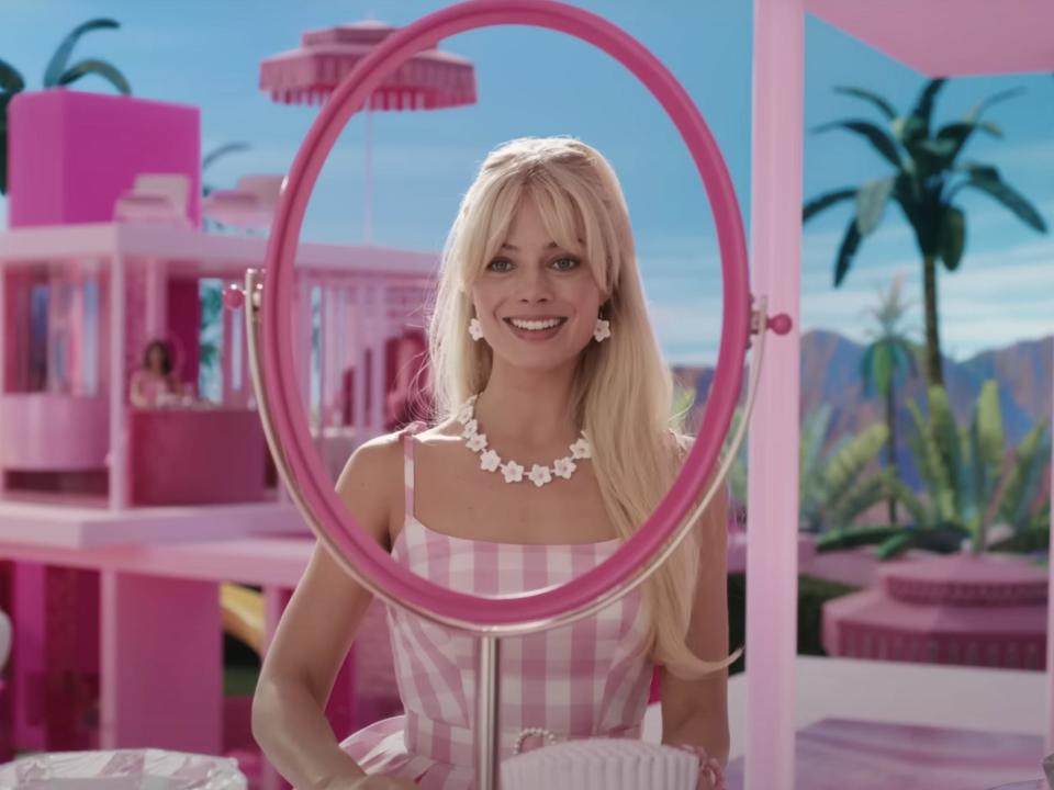 A screenshot from the "Barbie" trailer where Margot Robbie is looking into a pink mirror while standing in a pink apartment which looks out over a tropical-looking landscape with palm trees and mountains.