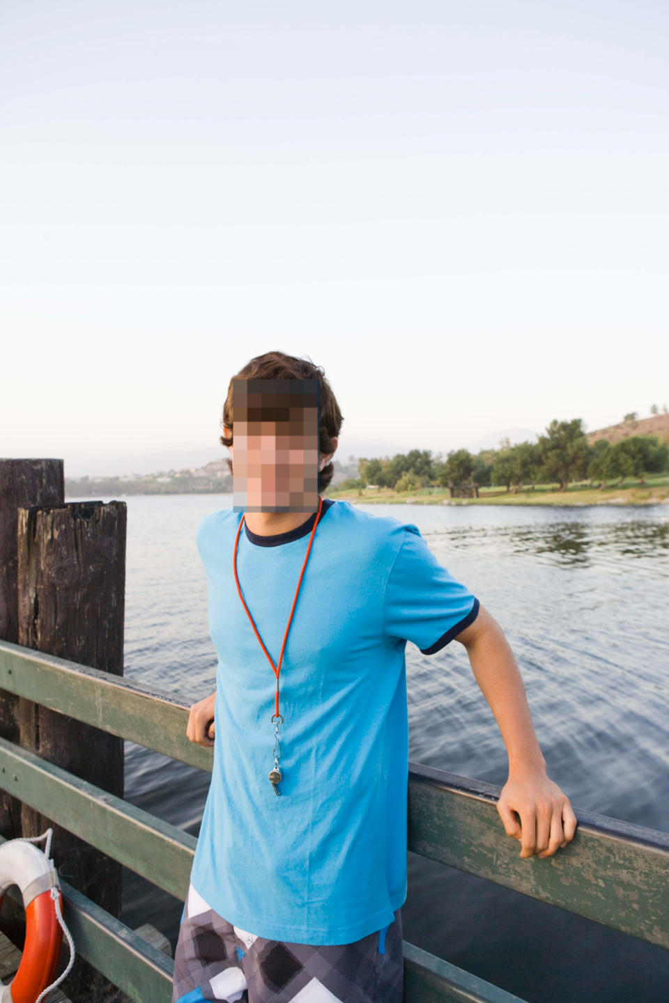 A young man, smiling, stands by a lakeside fence wearing a blue shirt with a whistle necklace. Trees and water are in the background