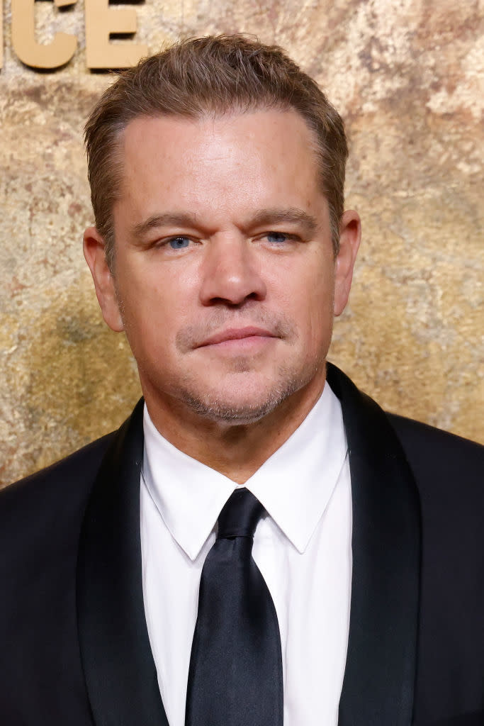 Matt Damon in a formal black suit and tie, posing at an event with a golden textured background