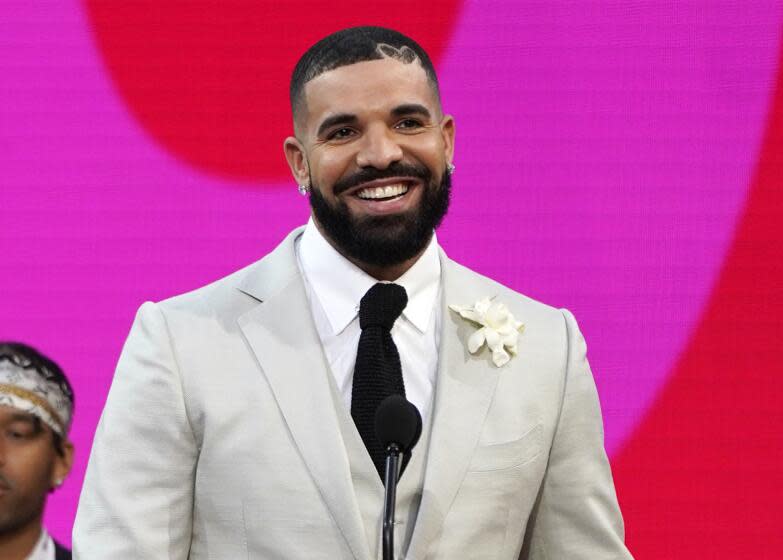 Drake smiles in front of a microphone while wearing a light gray suit and black tie.