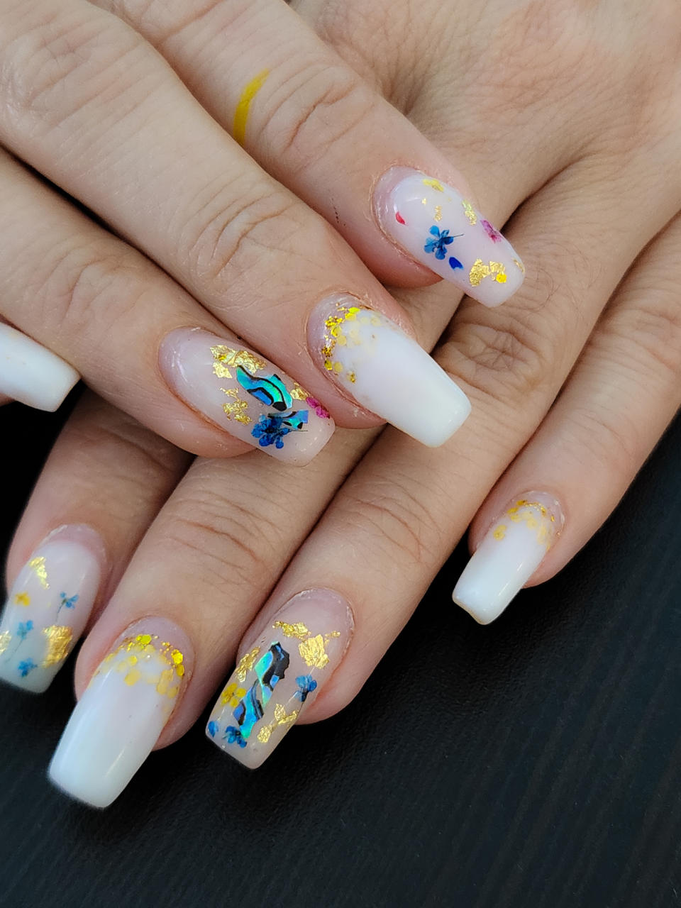 A close-up of hands with long, white-tipped acrylic nails featuring blue floral designs and gold foil accents