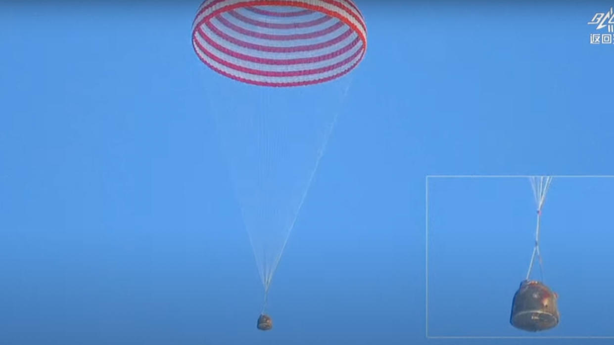  A dark-brown capsule descends through a blue sky beneath a red and white striped parachute. 