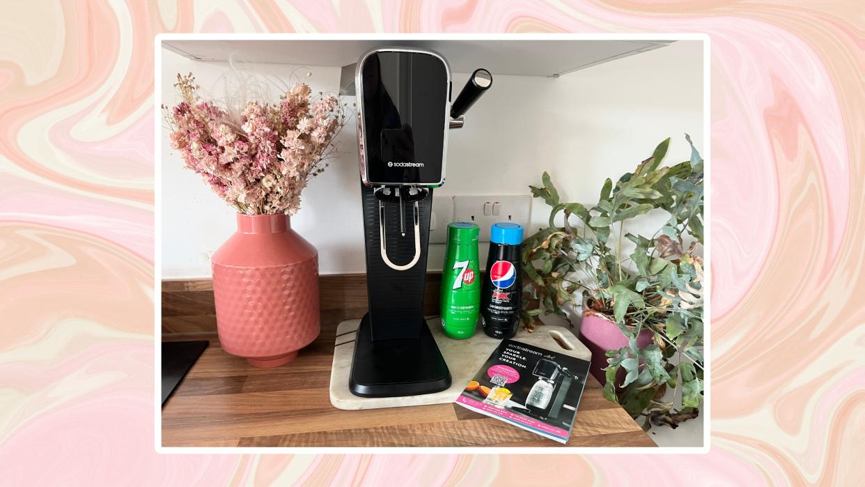  A pink swirl background with a photo of a black SodaStream Art on a marble board by a vase of dried flowers 