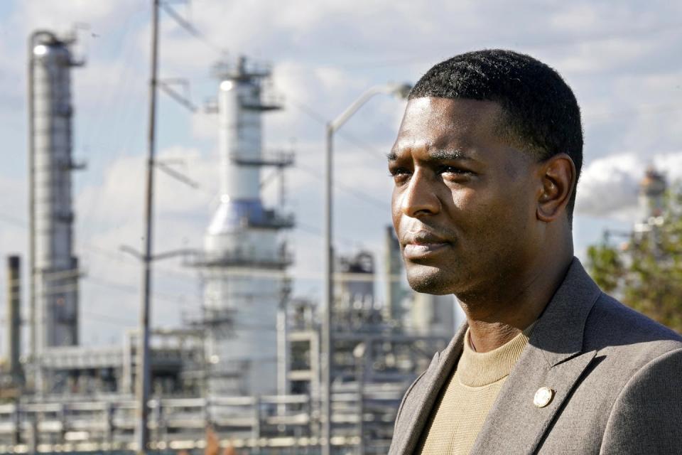 A man in a sport coat stands in front of a refinery.