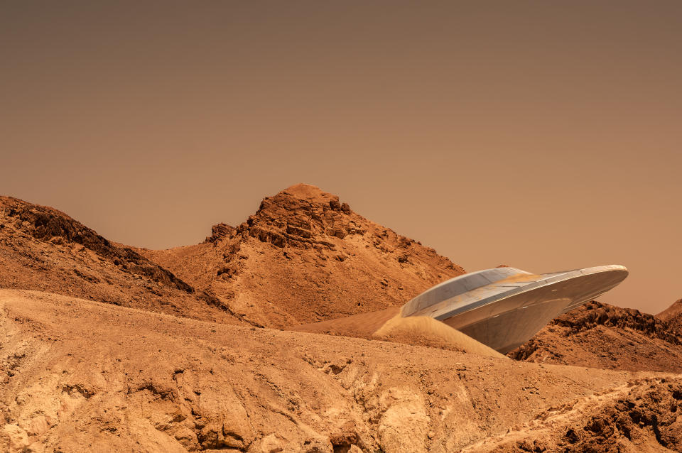 A sleek, futuristic spacecraft is partially buried in a desert landscape with rocky hills in the background. No people are present