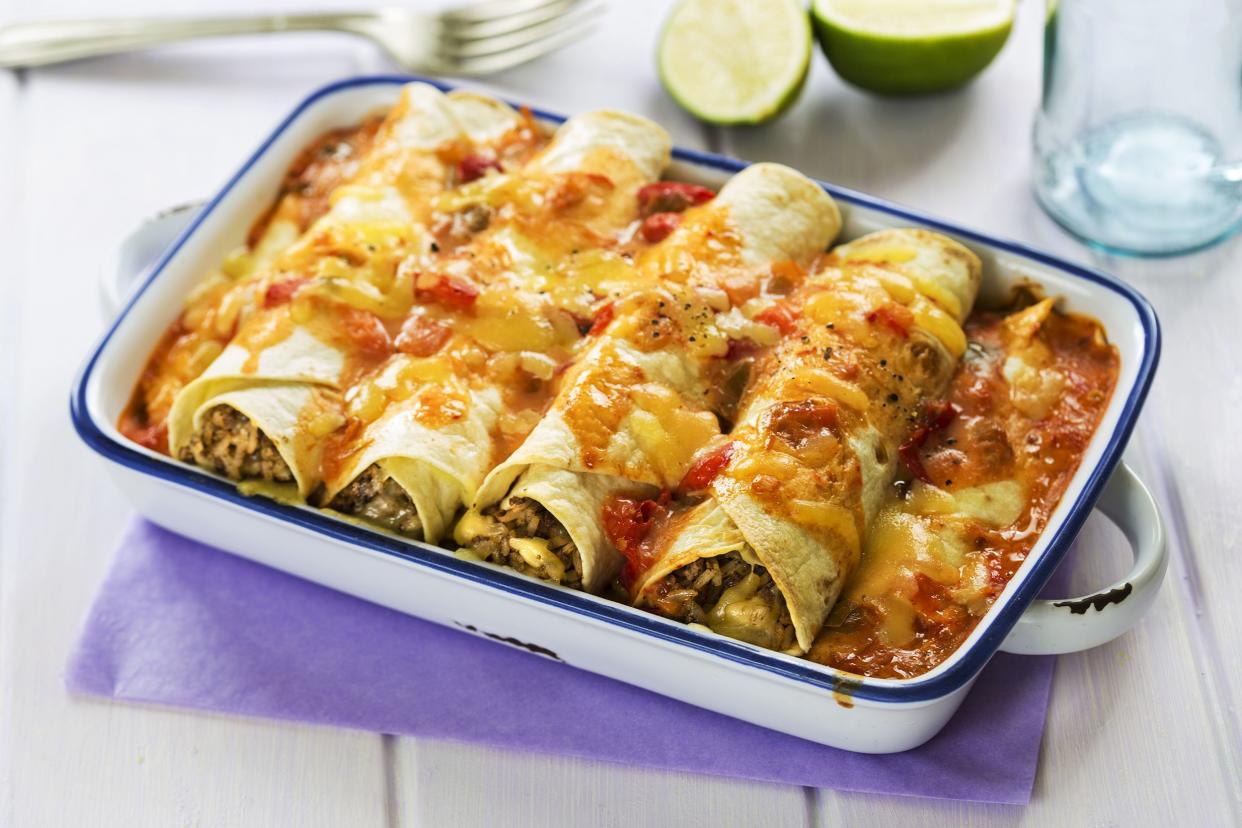 Four microwave beef enchiladas in a blue-lined white enamel gratin dish, on a lavender-colored cloth on a white wooden table, surrounded by a fork and a cut lime, both blurred