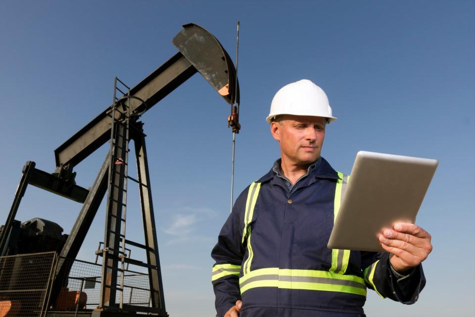 A worker holds a tablet while standing in front of an oil rig.